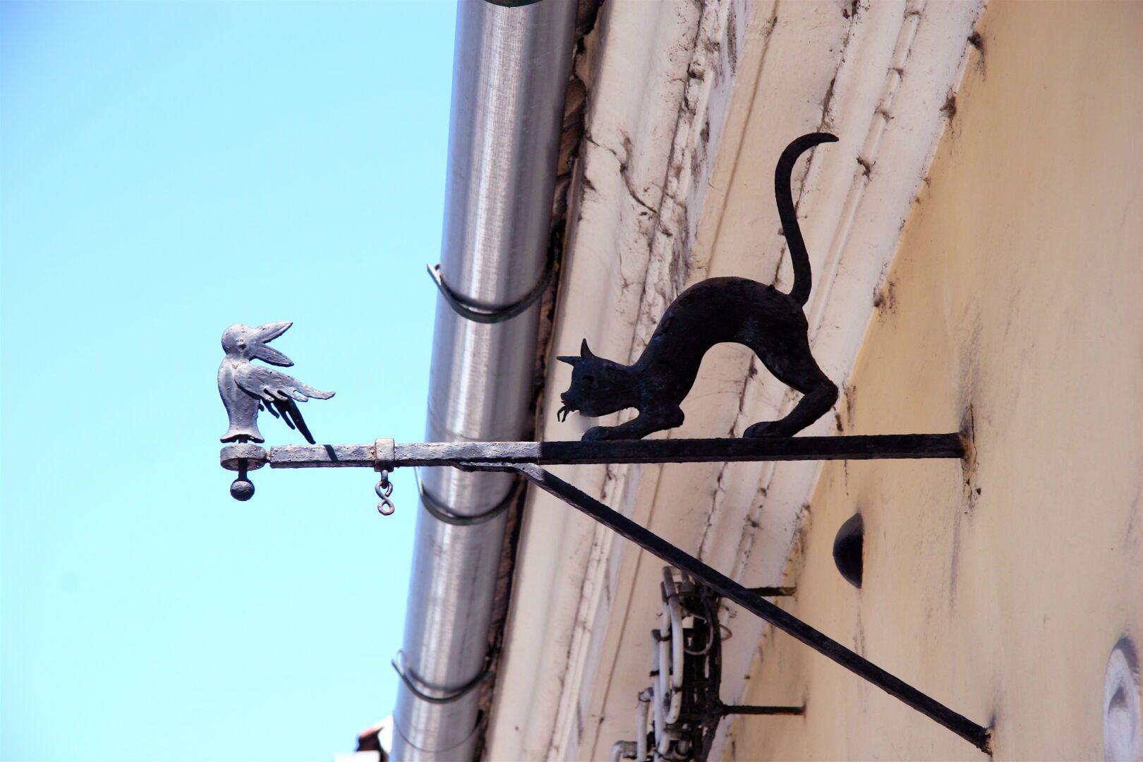 Cast iron sign support on a wall, with a silver-painted bird at the end and the silhouette of a black-painted cat about to pounce from the shadows.