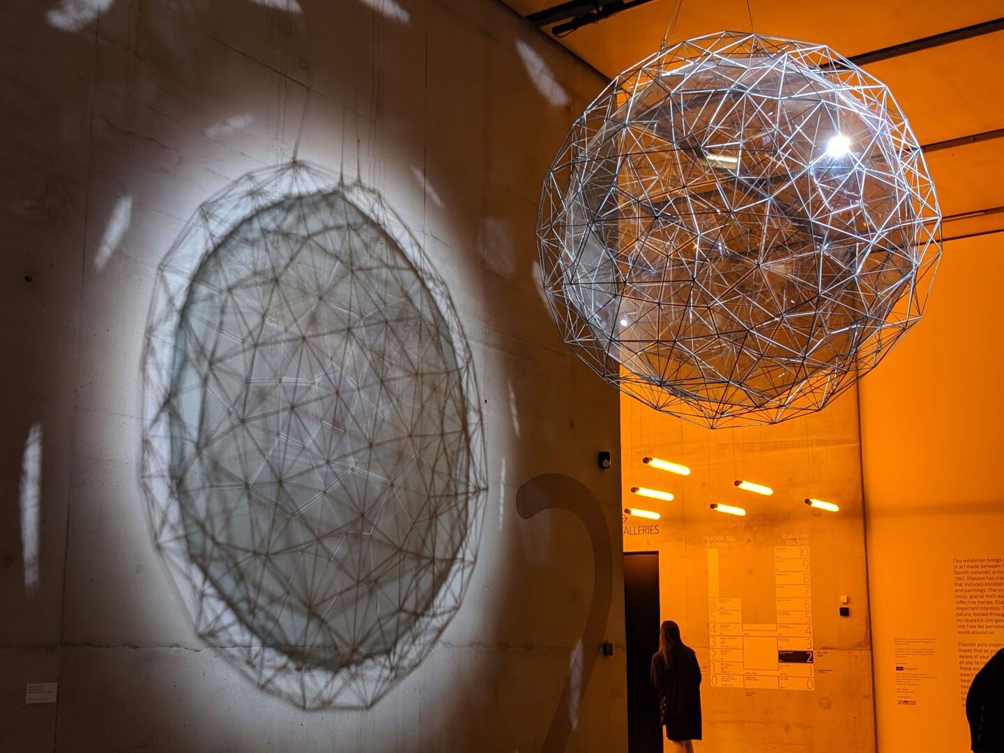 A concrete wall shows a remarkably 3D shadow of a sphere, resulting from the projection of light throufgh a wire geodesic globe containing a grey glass sphere that is suspended in front of the wall. The shadow is more striking than the globe, raising the question of which is the artwork. The background is a rich sodium orange from another exhibit