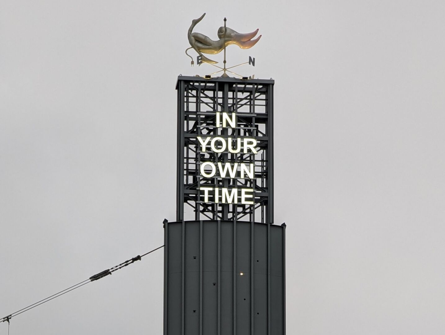 Top portion of sculpture at Brussels South Station. Topped by a gold weathervane, a black support structure on a ribbed black obelisk has the text "In Your Own Time"